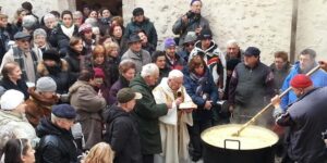 Crowd of local residents watching a priest bless a large pot of food outdoors.